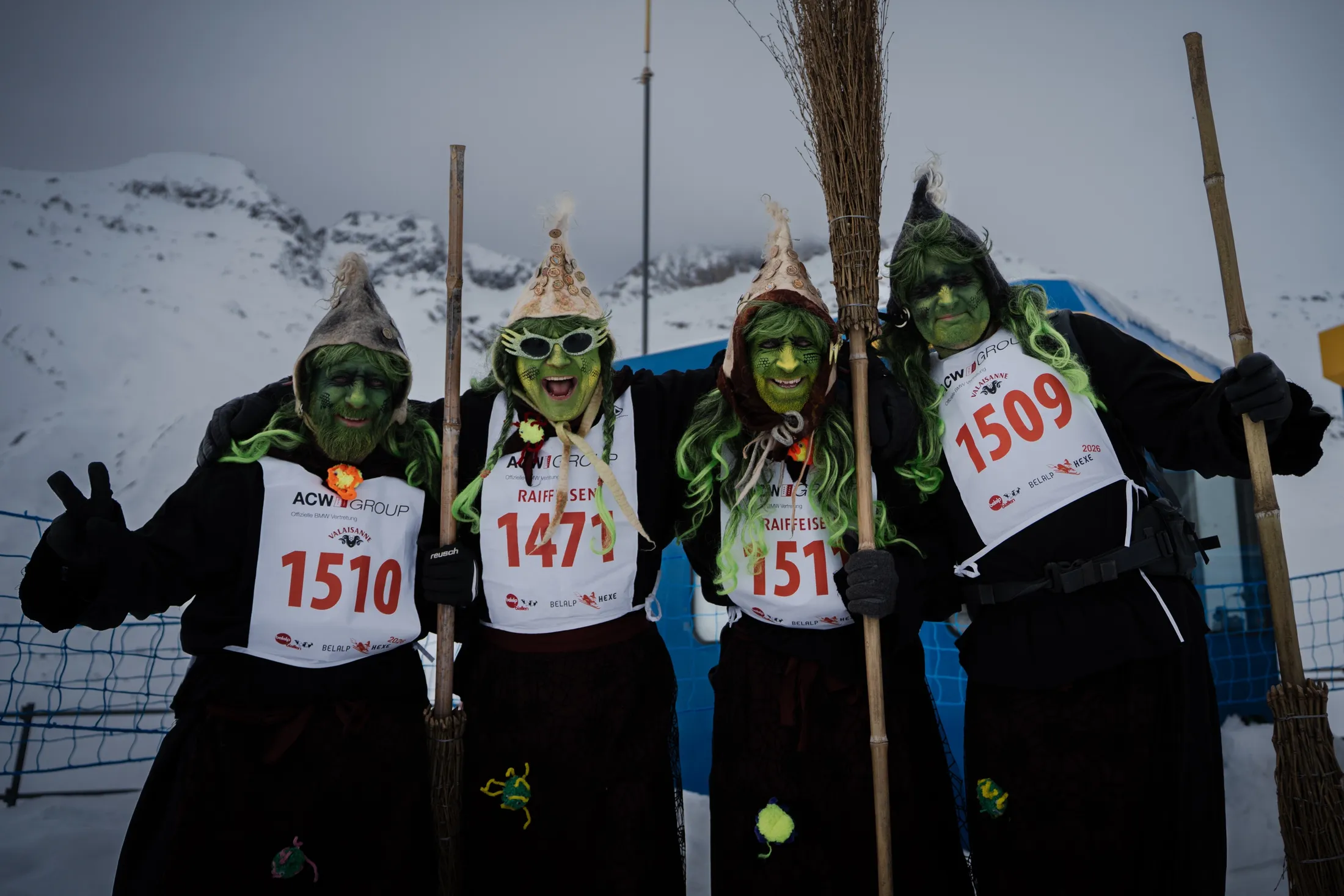 Vier grün geschminkte Hexen mit Startnummern und Besen posieren vor der verschneiten Belalp