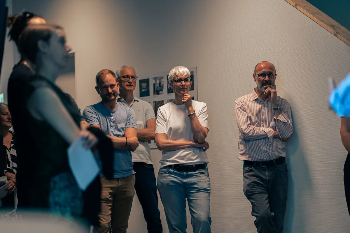 Gäste stehen der Wand entlang und hören einer Präsentation zu