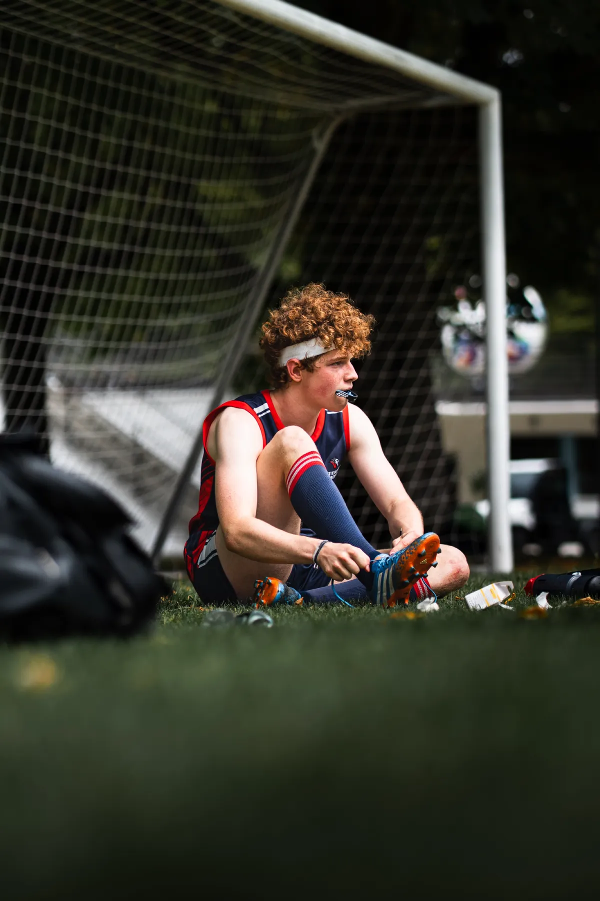 Junger Spieler mit roten Locken, Stirnband und Mundschutz sitzt vor dem Tor im Gras und schnürt seinen Schuh