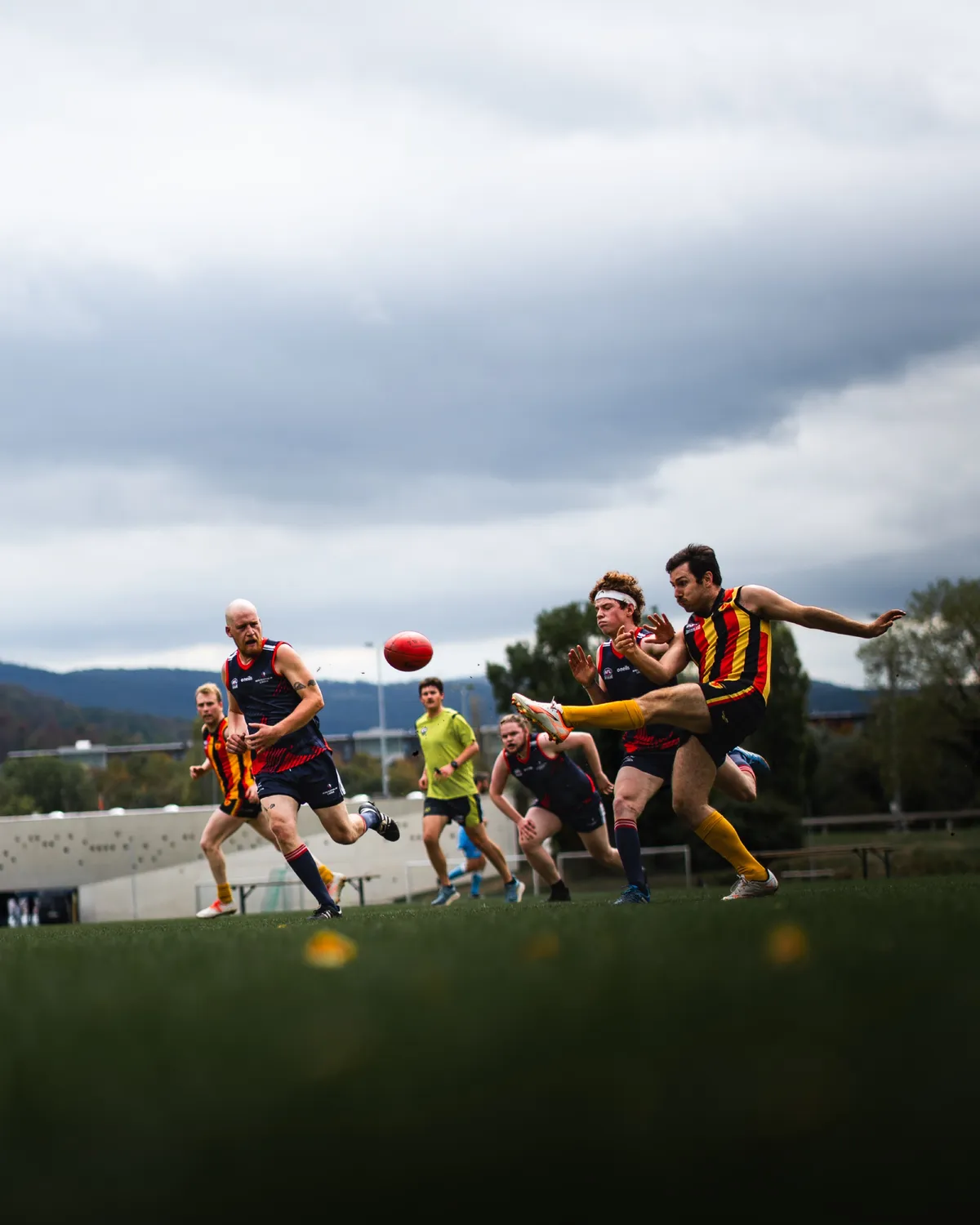 Spieler im Streifentrikot kickt den Ball, mehrere Gegenspieler und der Schiedsrichter sprinten heran, im Hintergrund Wolken und Hügel