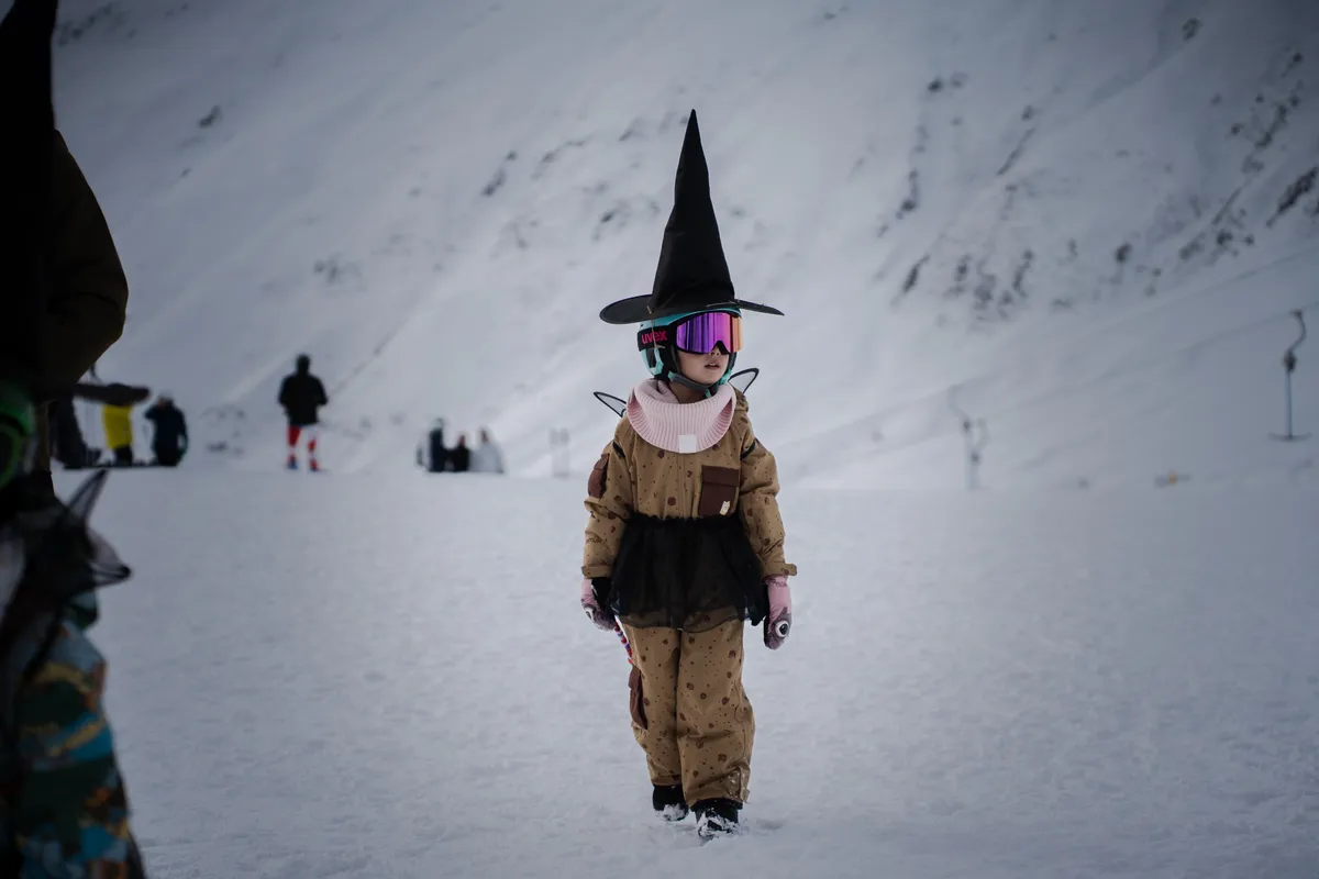Kind im Hexenkostüm mit spitzem Hut und violetter Skibrille steht im Schnee