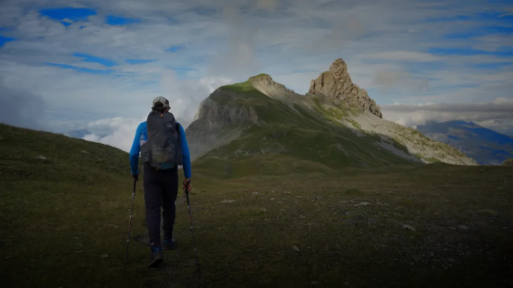 Filmstill: Wanderer mit Rucksack und Stöcken unterwegs Richtung Lobhörner, der felsige Gipfel ragt aus dem grünen Grat