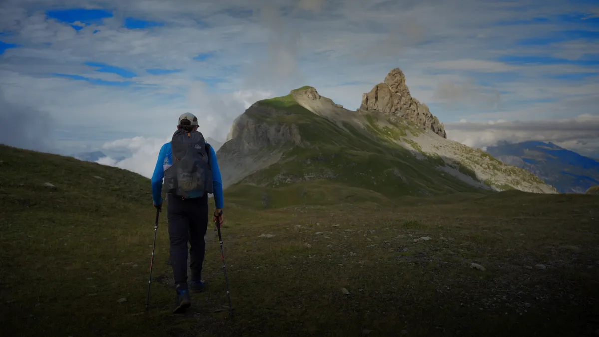 Filmstill: Wanderer mit Rucksack und Stöcken unterwegs Richtung Lobhörner, der felsige Gipfel ragt aus dem grünen Grat