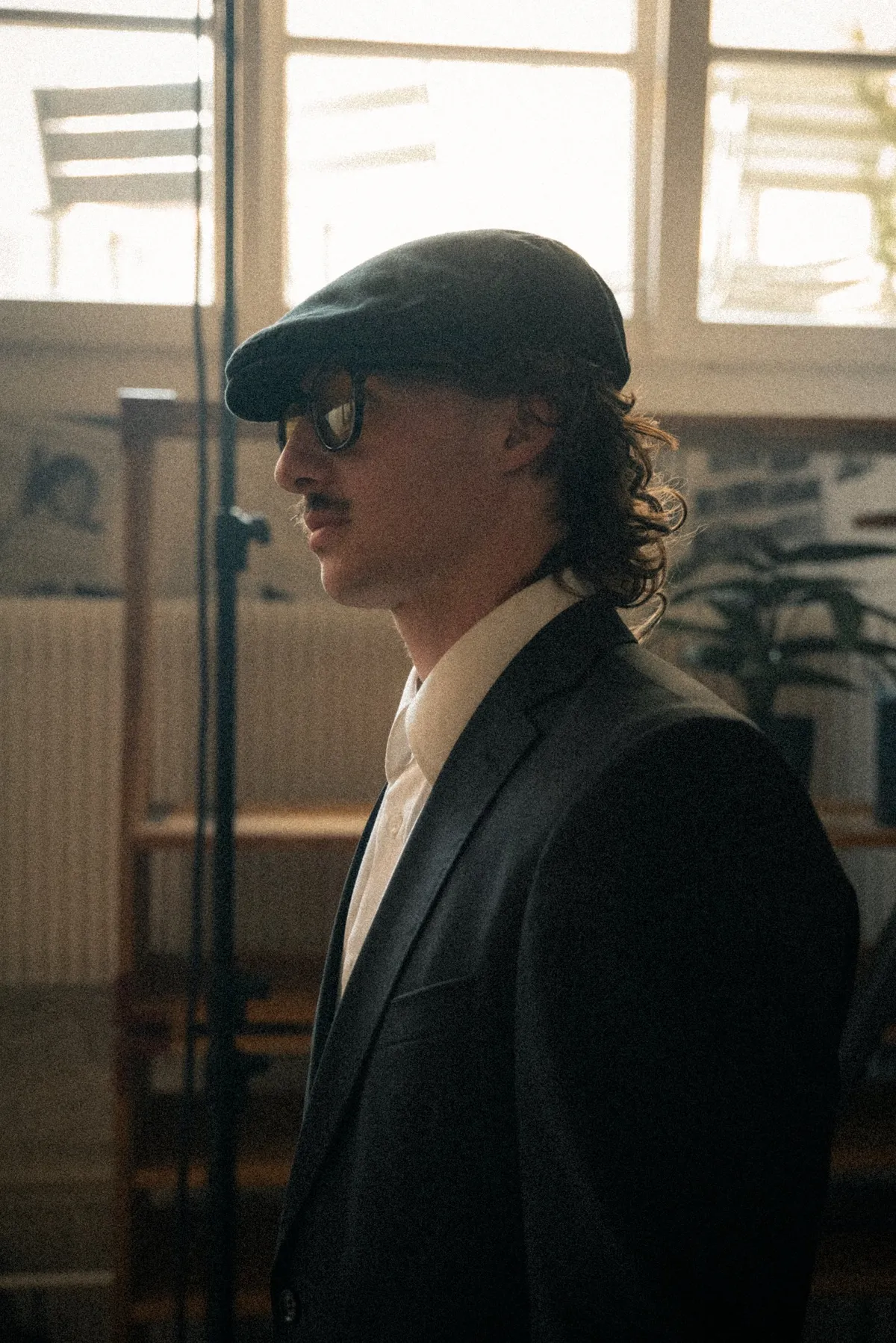 Maël mit Mütze, Sonnenbrille und Anzug vor einem hellen Fenster