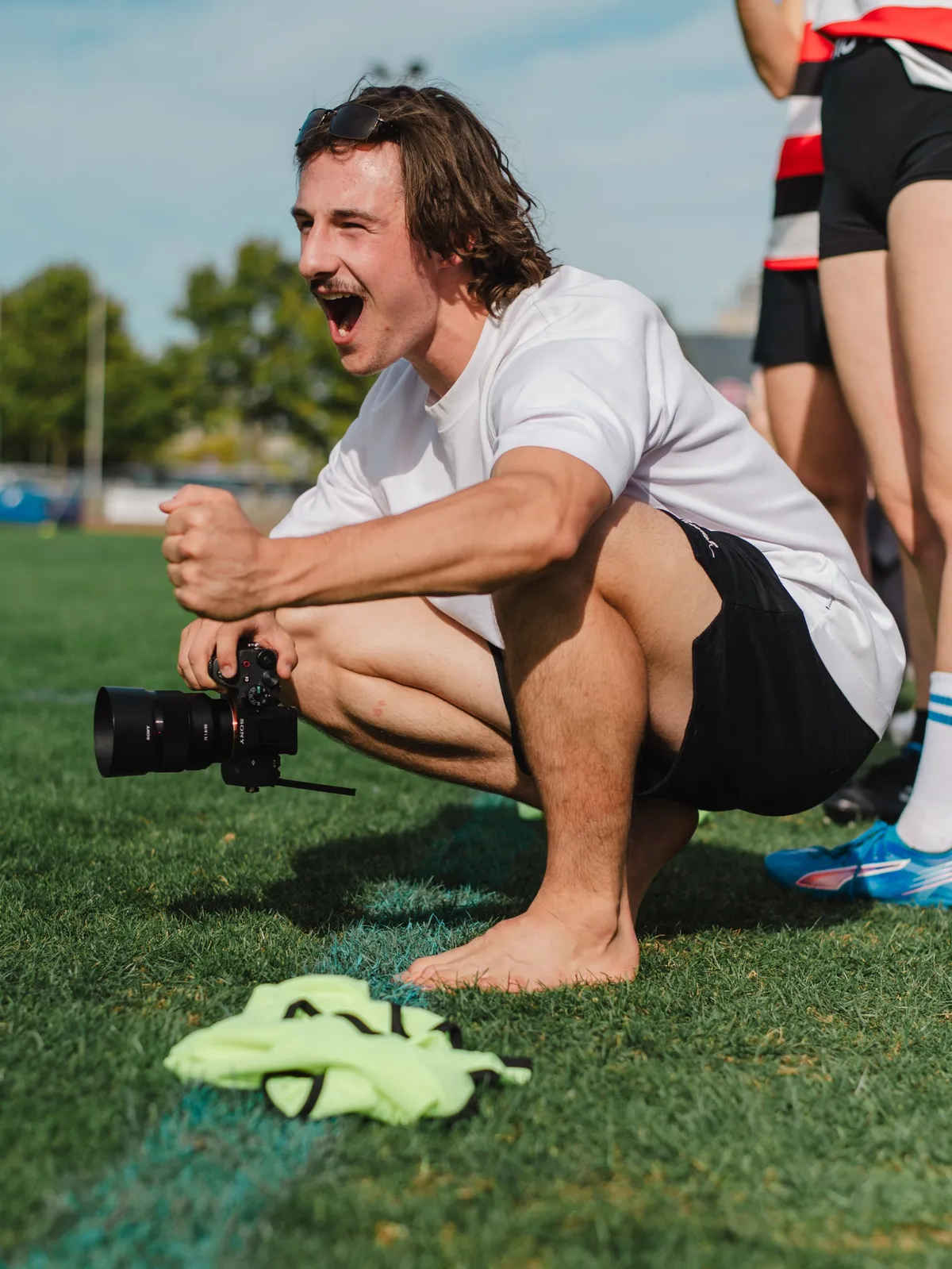 Maël kauert lachend mit der Kamera am Rand eines Fussballplatzes
